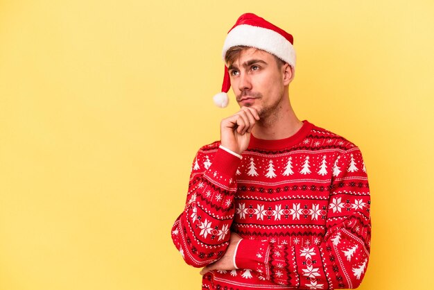 Young caucasian man celebrating Christmas isolated on yellow background looking sideways with doubtful and skeptical expression.