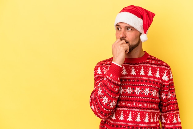 黄色の背景に分離されたクリスマスを祝う若い白人男性は、コピースペースを見ている何かについて考えてリラックスしました。