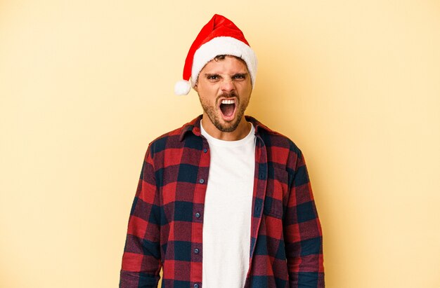 young caucasian man celebrating christmas isolated on yellow background screaming very angry and aggressive.