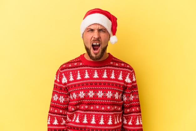 young caucasian man celebrating christmas isolated on yellow background screaming very angry and aggressive.