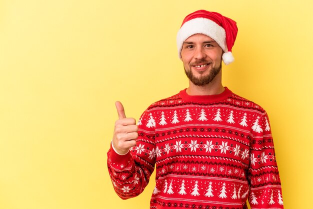 Young caucasian man celebrating Christmas isolated on yellow background smiling and raising thumb up