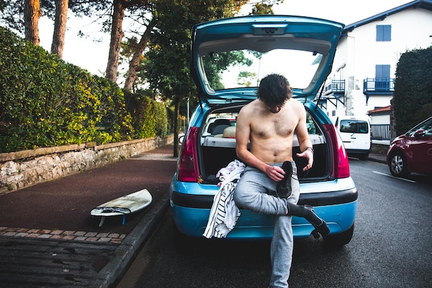 Молодой кавказский мужчина переодевается в багажнике автомобиля после серфинга на пляже.