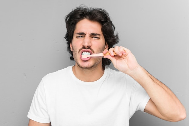 Young caucasian man cleaning his teeth with a toothbrush isolated in a grey wall