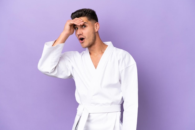 Young caucasian man doing karate isolated on purple background doing surprise gesture while looking to the side