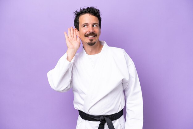 Foto giovane uomo caucasico che fa karate isolato su sfondo viola ascoltando qualcosa mettendo la mano sull'orecchio