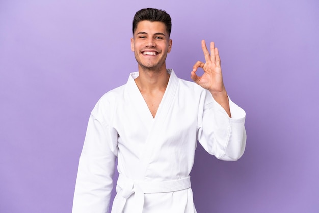 Young caucasian man doing karate isolated on purple background showing ok sign with fingers