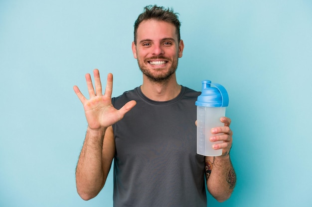 Young caucasian man drinking milkshake isolated on  blue background smiling cheerful showing number five with fingers.