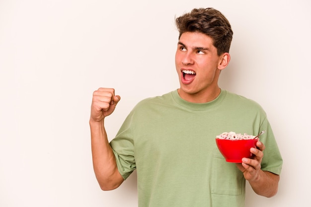 Young caucasian man eating cereals isolated on white background raising fist after a victory winner concept