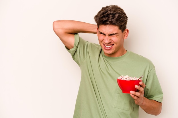 Young caucasian man eating cereals isolated on white background touching back of head thinking and making a choice