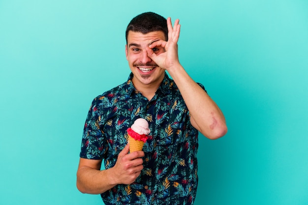 青い背景に分離されたアイスクリームを食べている若い白人男性は、目で大丈夫なジェスチャーを続けて興奮していました。
