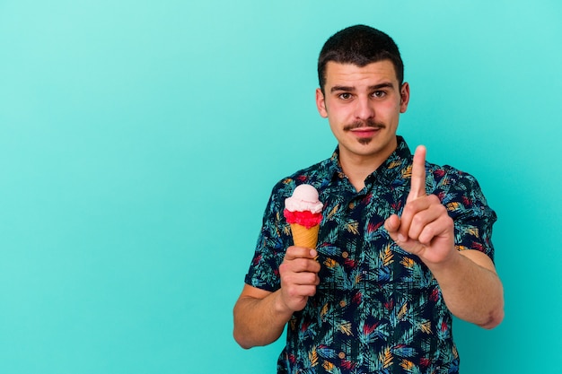 Young caucasian man eating an ice cream isolated on blue wall showing number one with finger.