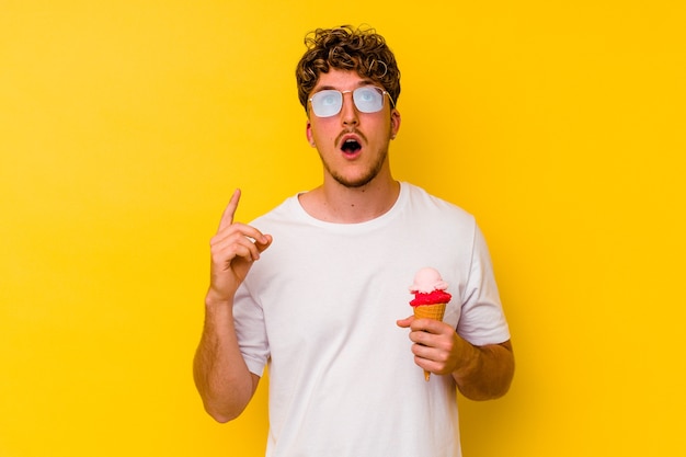 Young caucasian man eating an ice cream isolated on yellow background pointing upside with opened mouth.