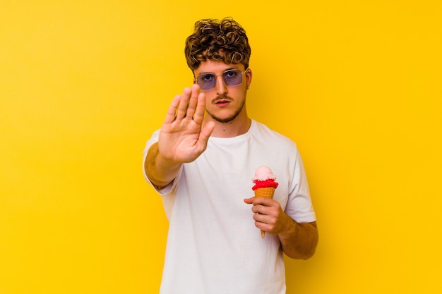 Young caucasian man eating an ice cream isolated on yellow wall standing with outstretched hand showing stop sign, preventing you.