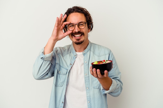 Young caucasian man eating a ramen isolated on white excited keeping ok gesture on eye.