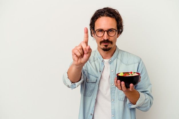 Giovane uomo caucasico che mangia un ramen isolato su bianco che mostra il numero uno con il dito.