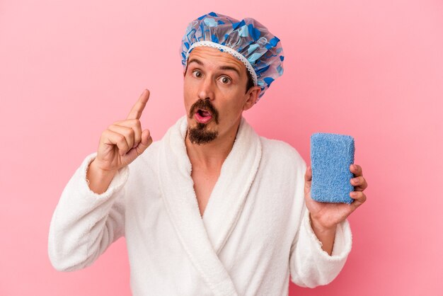 Young caucasian man going to the shower holding sponge isolated on pink background having an idea, inspiration concept.