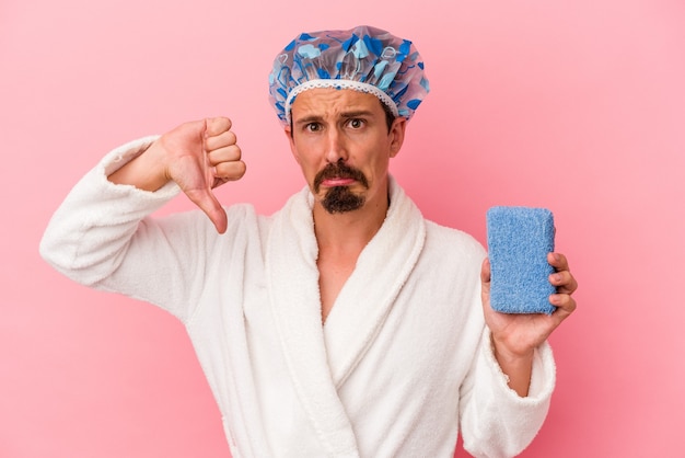 Young caucasian man going to the shower holding sponge isolated on pink background showing a dislike gesture, thumbs down. Disagreement concept.