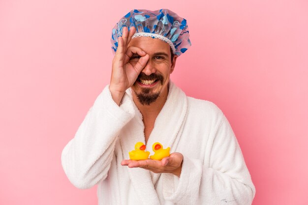 Young caucasian man going to the shower with rubber ducks isolated on pink background excited keeping ok gesture on eye.