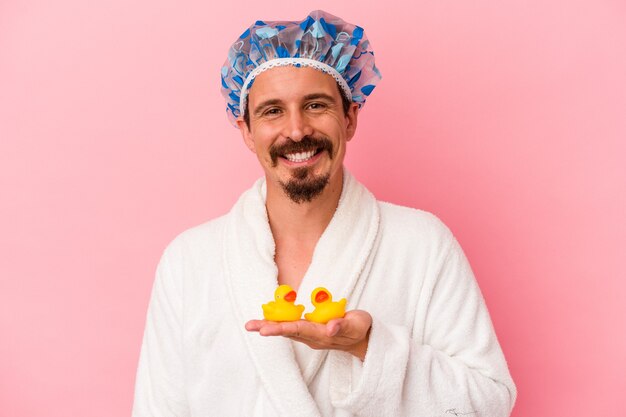 Young caucasian man going to the shower with rubber ducks isolated on pink background happy, smiling and cheerful.