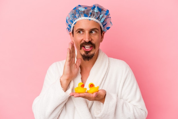 Young caucasian man going to the shower with rubber ducks\
isolated on pink background is saying a secret hot braking news and\
looking aside