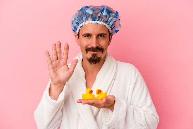 Молодой кавказский человек собирается в душ с резиновыми утками, изолированными на розовом фоне, улыбаясь, веселый, показывая номер пять с пальцами.
