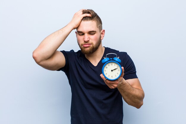 Young caucasian man holding alarm clock being shocked, she has remembered important meeting.