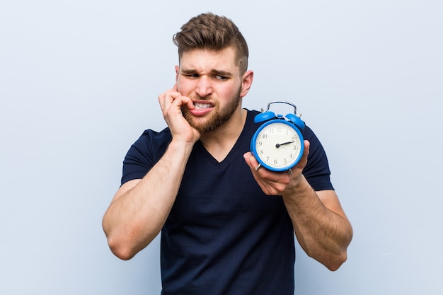 Young caucasian man holding alarm clock biting fingernails, nervous and very anxious.