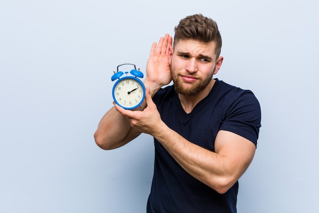 Young caucasian man holding alarm clock trying to listening a gossip.