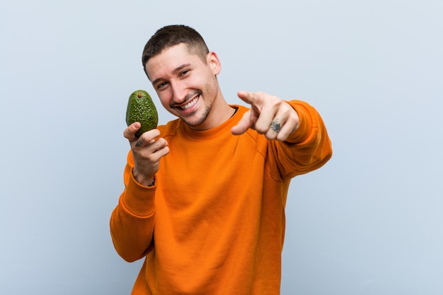 Giovane uomo caucasico che tiene un sorriso allegro di un avocado che indica la parte anteriore.