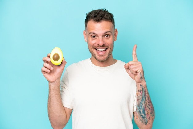 Giovane uomo caucasico che tiene un avocado isolato su sfondo blu che indica una grande idea