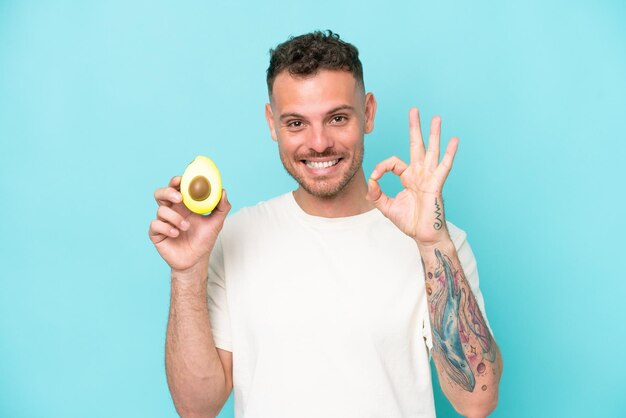 Молодой кавказский мужчина держит авокадо на синем фоне, показывая пальцами знак "ок"
