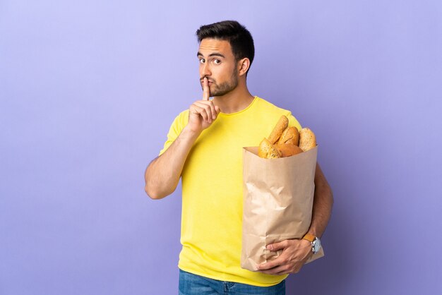 Giovane uomo caucasico che tiene una borsa piena di pane isolato su sfondo viola facendo gesto di silenzio