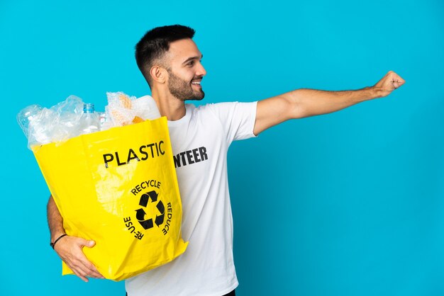 Young caucasian man holding a bag full of plastic bottles to recycle isolated on blue background giving a thumbs up gesture