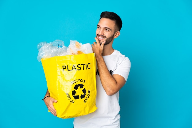 Young caucasian man holding a bag full of plastic bottles to recycle isolated on blue background looking to the side and smiling