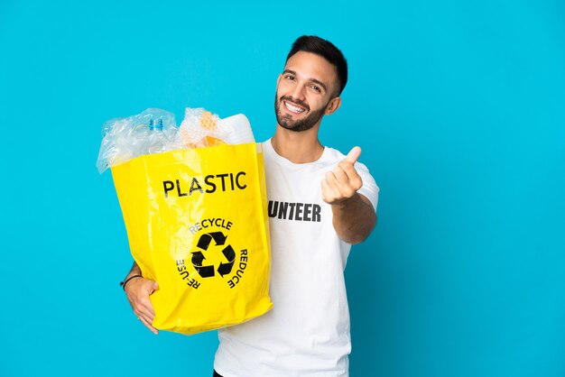 Young caucasian man holding a bag full of plastic bottles to recycle isolated on blue background making money gesture