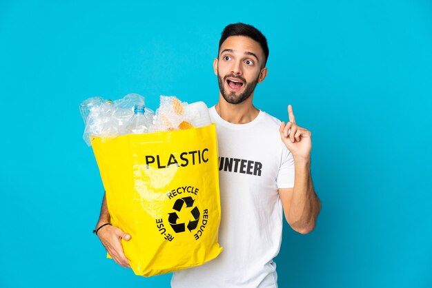 Young caucasian man holding a bag full of plastic bottles to recycle isolated on blue background thinking an idea pointing the finger up