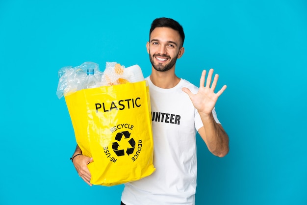 Young caucasian man holding a bag full of plastic bottles to recycle isolated on blue wall counting five with fingers
