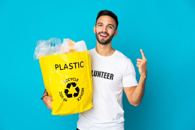 Young caucasian man holding a bag full of plastic bottles to recycle isolated on blue wall showing and lifting a finger in sign of the best