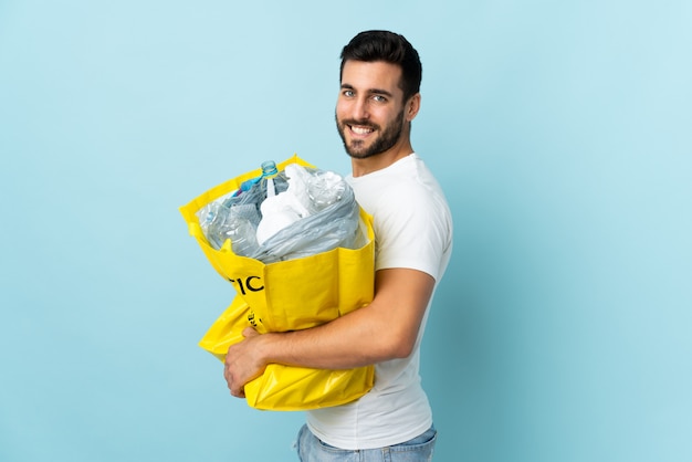 Young caucasian man holding a bag full of plastic bottles to recycle isolated on blue wall smiling a lot