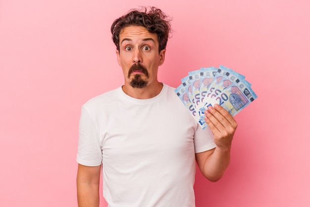 Young caucasian man holding banknotes isolated on pink background shrugs shoulders and open eyes confused.
