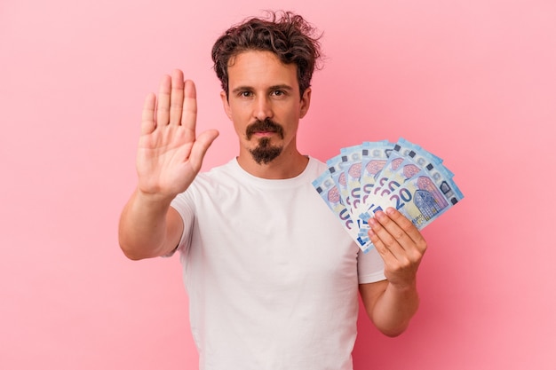 Young caucasian man holding banknotes isolated on pink background standing with outstretched hand showing stop sign, preventing you.