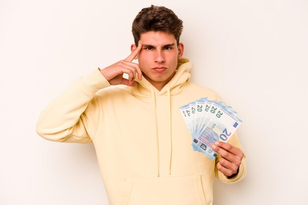 Young caucasian man holding banknotes isolated on white background pointing temple with finger thinking focused on a task