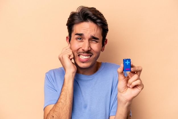 Young caucasian man holding a batterie isolated on beige background covering ears with hands