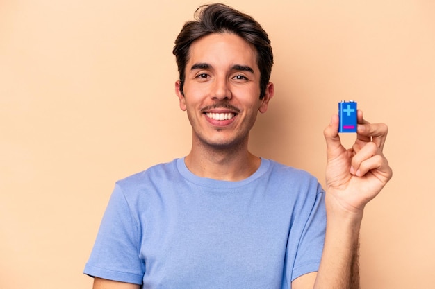 Foto giovane uomo caucasico in possesso di una batteria isolata su sfondo beige felice, sorridente e allegro.