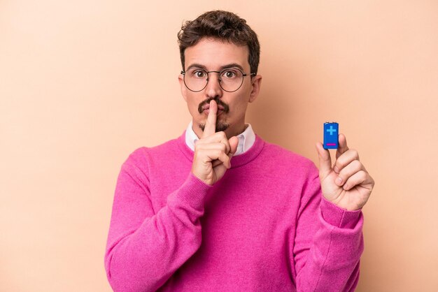 Young caucasian man holding batteries isolated on beige background keeping a secret or asking for silence