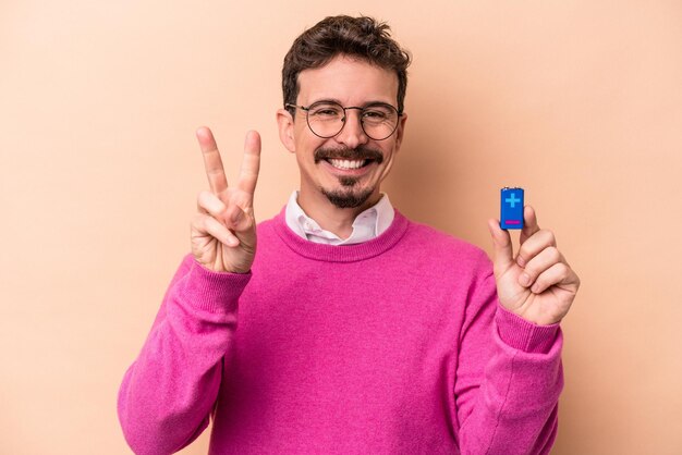 指で2番目を示すベージュの背景に分離された電池を保持している若い白人男性。
