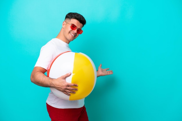 Young caucasian man holding a beach ball isolated on blue background extending hands to the side for inviting to come