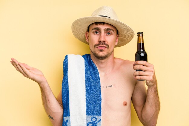 Young caucasian man holding a beer isolated on yellow background