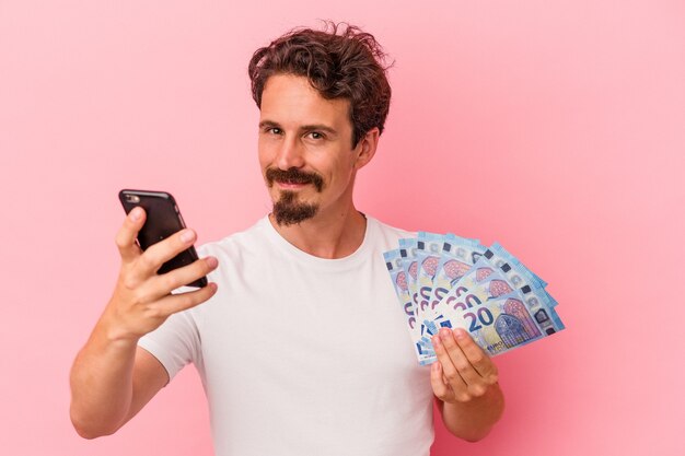 Young caucasian man holding bills and piggy bank isolated on pink background