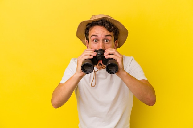 Young caucasian man holding binoculars isolated on yellow background